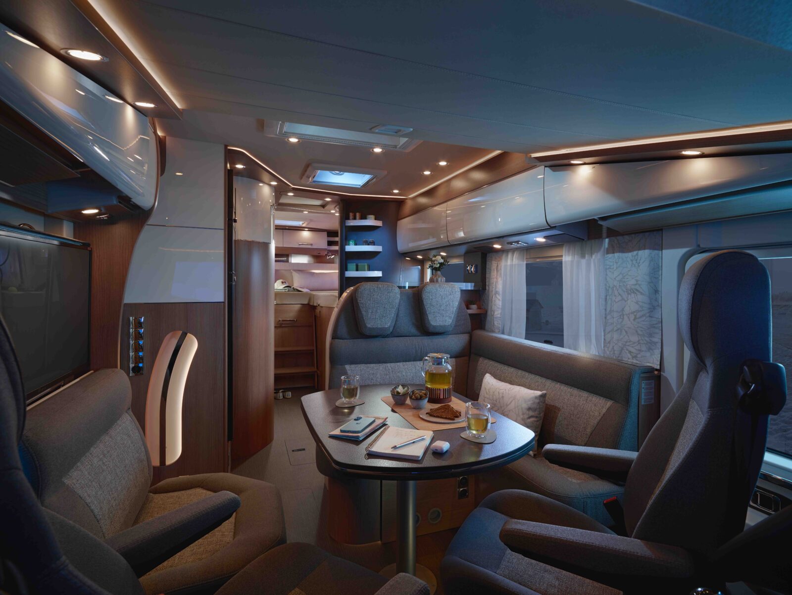 Ein modernes Wohnmobil-Interieur mit bequemen Sitzgelegenheiten um einen Esstisch, Getränken und Snacks auf dem Tisch, stimmungsvoller Beleuchtung und großen Fenstern mit Blick nach draußen. Im Hintergrund sind Regale und Schränke zu sehen.