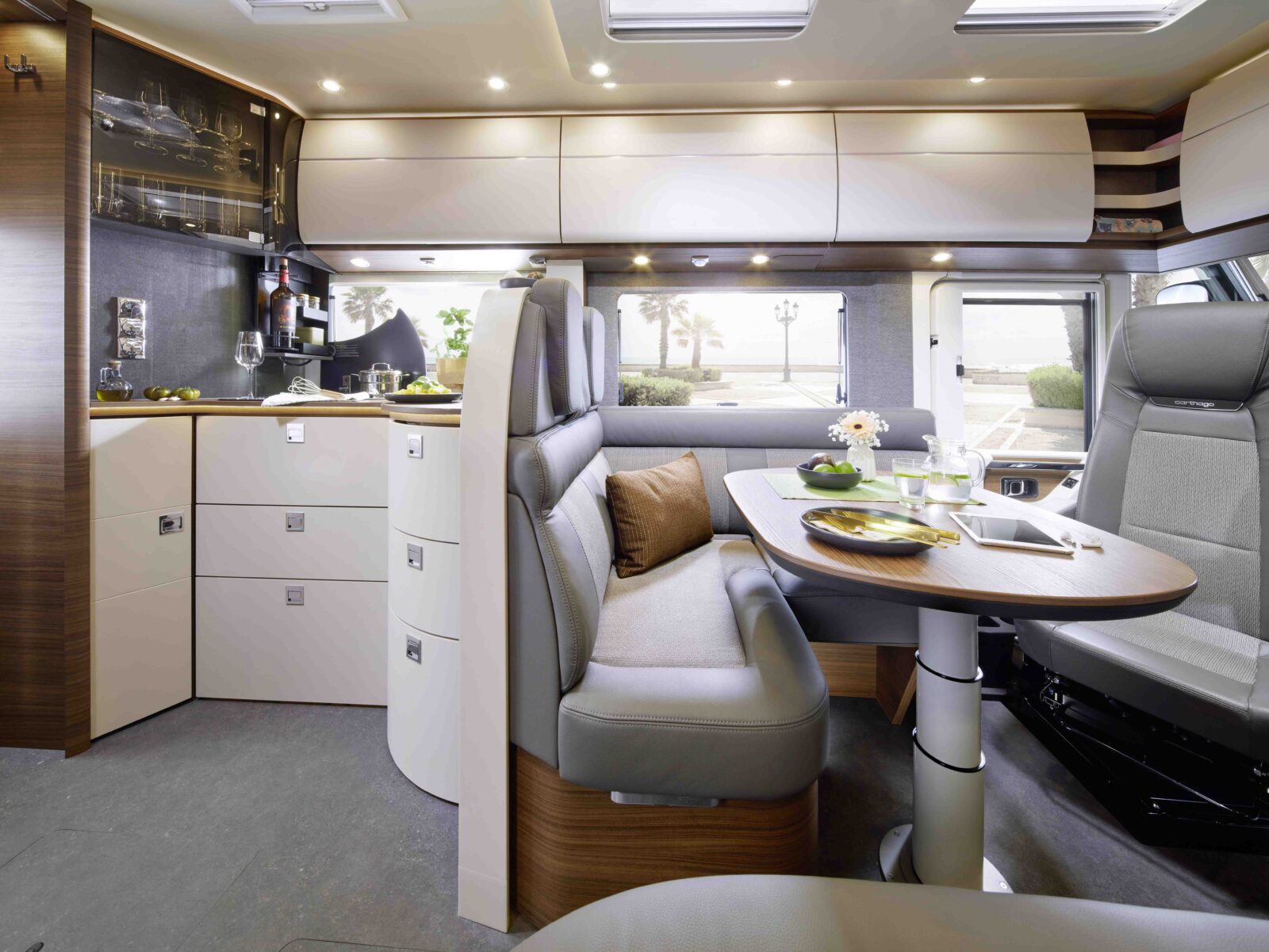 Modernes Wohnmobil-Interieur mit einer kompakten Küche, Regalen, einem Esstisch für zwei Personen und gepolsterten Sitzen. Große Fenster sorgen für natürliches Licht und einen Blick nach draußen. Das Design ist in hellem Holz und neutralen Tönen gehalten.