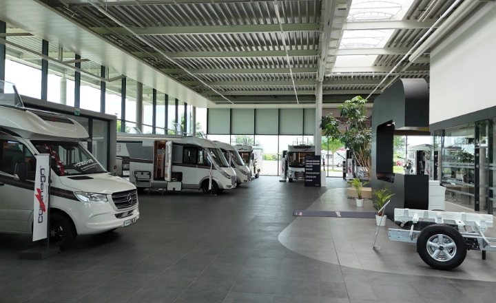 Ein heller, geräumiger Ausstellungsraum mit mehreren vollintegrierten Wohnmobil- und teilintegrierten Wohnmobil-Modellen, großen Fenstern, die viel Tageslicht hereinlassen, und einem Wohnwagen im Vordergrund. Das moderne Interieur ist durchweg mit Topfpflanzen bepflanzt.