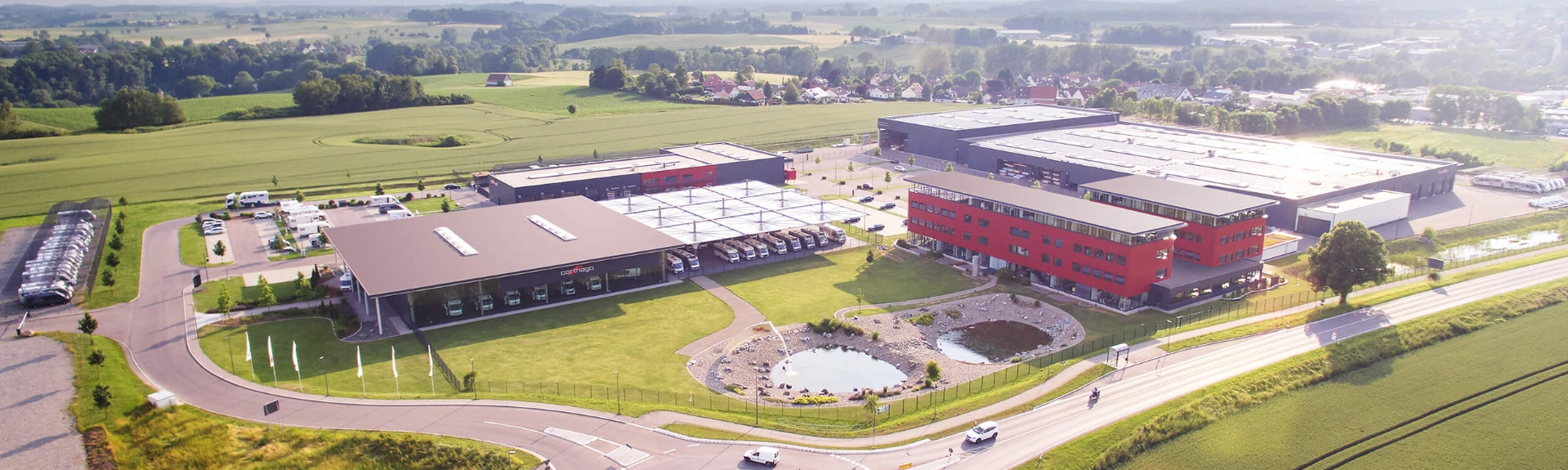 Luftaufnahme eines modernen Industriekomplexes mit roten und grauen Gebäuden, Parkplätzen mit einem reisemobil und einem vollintegrierten wohnmobil sowie einem kleinen Teich, umgeben von grünen Feldern und Bäumen unter einem klaren Himmel. Eine Straße führt entlang des Komplexes.