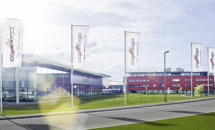 Ein moderner Gebäudekomplex mit großen Glasfenstern und roter Fassade steht hinter hohen weißen Fahnen mit dem Carthago-Logo und -Slogan, die an einem sonnigen Tag auf das hochwertige integrierte Wohnmobil hinweisen, wobei die Sonne im Vordergrund flackert.