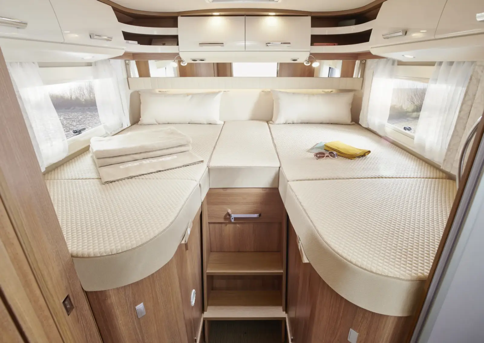 Das gemütliche Wohnmobil-Schlafzimmer im Carthago C2-Tourer verfügt über ein großes Bett mit beiger Bettwäsche, gefaltete Handtücher und eine Sonnenbrille, die auf einem Buch liegt. Holzschränke, weiß getönte Fenster und eingebaute Stauräume sorgen für Komfort und Stil.