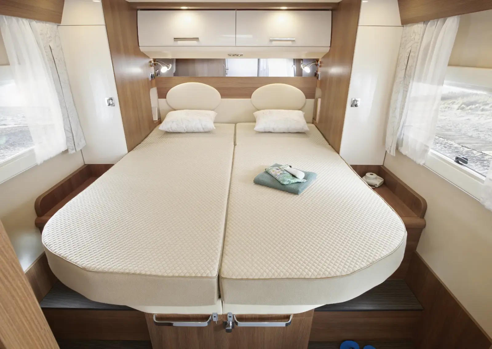 Ein ordentlich gemachtes Doppelbett mit cremefarbenem Bettzeug und zwei Kissen steht in einem modernen Carthago c2-Wohnmobil, umgeben von Holzvertäfelung und Fenstern mit weißen Vorhängen. Ein gefaltetes Handtuch und Toilettenartikel liegen auf dem Bett.