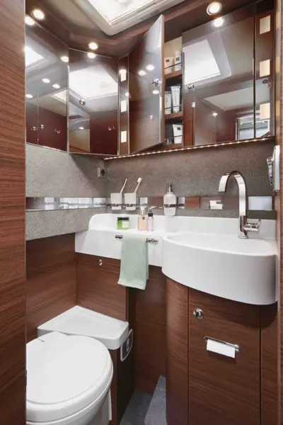 Modernes, kompaktes Badezimmer der Carthago Chic C Line mit holzgetäfelten Schränken, weißem Waschbecken, Toilette, Spiegelschränken, Toilettenartikeln und sanfter Beleuchtung. Über dem Waschbecken hängt ein grünes Handtuch, und es gibt Ablagen für Körperpflegeartikel.