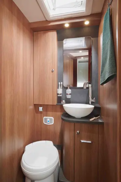 Kompaktes Badezimmer im Carthago C Tourer Teilintegriert mit holzgetäfelten Wänden, einer weißen Toilette, einem runden Waschbecken auf einer schwarzen Arbeitsplatte, einem rechteckigen Spiegel, kleinen Schränken, einem grünen Handtuch an der Wand und Deckenleuchten.