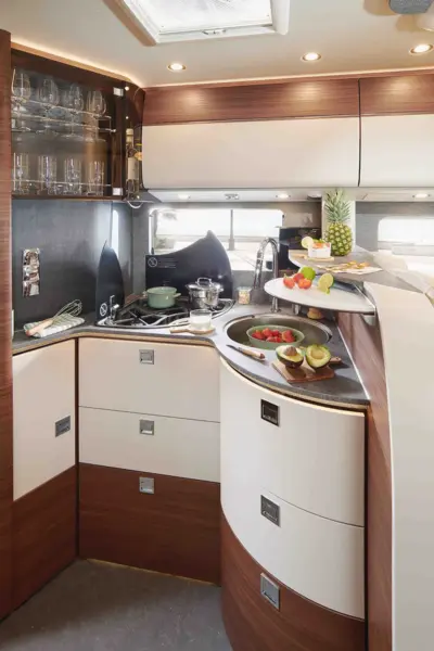 Eine moderne Wohnmobilküche im Carthago Chic C Line verfügt über geschwungene Holz- und weiße Schränke, eine Spüle, Obst und Gemüse auf der Theke, gestapelte Teller, Gläser in einem Schrank, ein Fenster und ein Oberlicht darüber, das natürliches Licht hereinlässt.