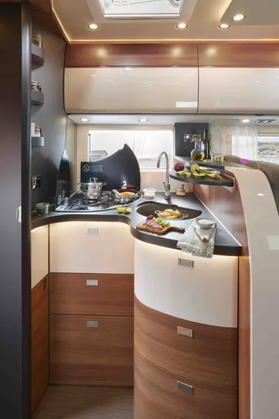 Moderne Wohnmobilküche in einem Carthago c tourer teilintegriert, mit geschwungenen Holzschränken, einer glatten Arbeitsplatte mit frischen Lebensmitteln, Edelstahlspüle, Herd mit Töpfen und Pfannen, Regale für Gewürze, Wein, Gemüse, und Unterschrankbeleuchtung.
