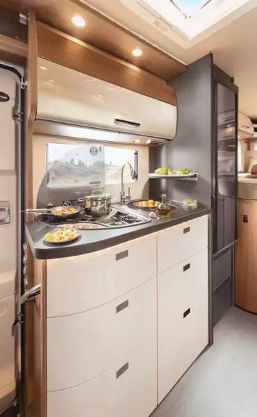 Moderne Wohnmobilküche im Carthago c tourer teilintegriert, mit glänzend weißen Schränken, einer grauen Arbeitsplatte, Herd, Spüle und frischen Lebensmitteln wie Salat und Brot. Natürliches Licht strömt durch ein Fenster mit Blick auf den Außenbereich.