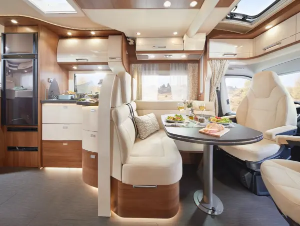 Der Innenraum des teilintegrierten Wohnmobils Carthago c tourer verfügt über einen gemütlichen Essbereich mit einem runden Tisch, cremefarbenen Sitzen, eleganten Holzschränken und großen Fenstern, die natürliches Licht hereinlassen.