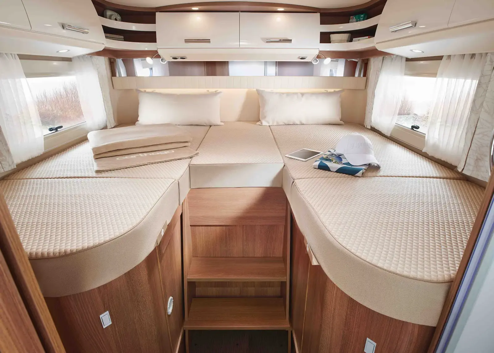 Innenraum eines Carthago C Tourer Teilintegrierten Wohnmobils mit zwei Einzelbetten, die durch eine Holztreppe getrennt sind, beigem Bettzeug, Kopfkissen und Hängeschränken. Auf einem Bett liegen ein Tablet und ein Hut. Große Fenster mit weißen Vorhängen lassen natürliches Licht herein.