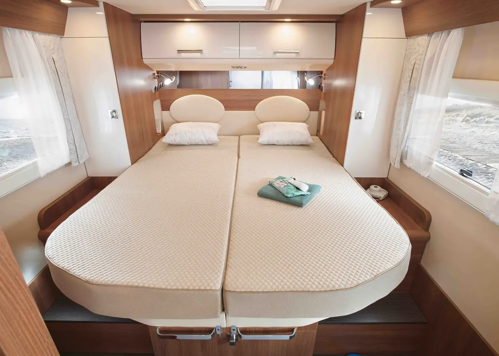 Ein ordentlich gemachtes Doppelbett mit beiger Bettwäsche und zwei Kissen steht in einem modernen Carthago C Tourer Teilintegrierten Wohnmobil, mit Fenstern auf beiden Seiten, weißen Vorhängen und gefalteten Handtüchern in der Mitte des Bettes.