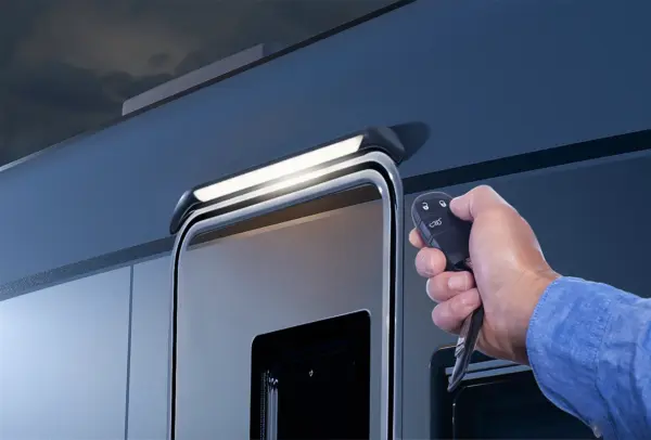 Eine Hand, die eine Fernbedienung und einen Schlüssel hält, zeigt auf die Tür eines modernen Carthago Liner for Two Wohnmobils bei Nacht, mit einer beleuchteten Lampe über der Tür.