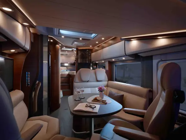 Luxuriöses Carthago Chic C Line Wohnmobil-Interieur mit cremefarbenen Ledersitzen, einem runden Tisch mit Weingläsern und einer Blumenvase, stimmungsvoller Beleuchtung und einem Bettbereich im Hintergrund. Die Atmosphäre ist gemütlich und einladend.