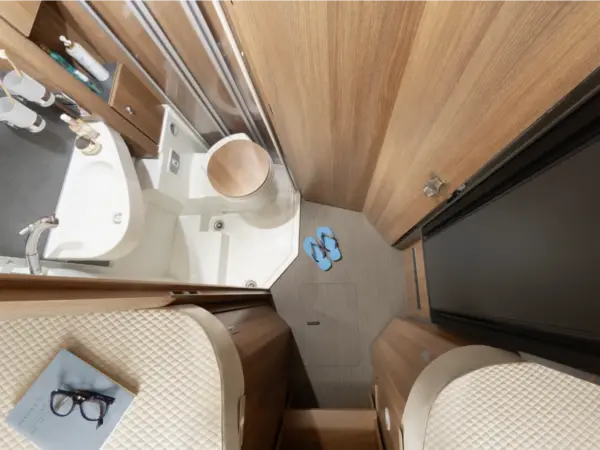 Ein kleines, modernes Badezimmer in einem Carthago C2-Tourer-Wohnmobil mit Holzwänden, einer kompakten Toilette und einem Waschbecken, blauen Flip-Flops auf dem Boden und einem Bett mit einem Buch und einer Brille daneben.