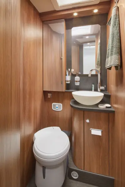 Das kompakte Badezimmer im Carthago C2-Tourer ist mit holzgetäfelten Wänden, einer modernen Toilette, einem runden Waschbecken auf einer schwarzen Arbeitsplatte, einem Wandspiegel, Regalen mit Toilettenartikeln, einem Hängetuch und einer hellen Deckenbeleuchtung ausgestattet.