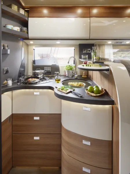 Moderne Wohnmobilküche im Carthago C2-Tourer mit Holz- und weißen Schränken, geschwungenen Arbeitsflächen, einem Gasherd mit Kochfunktion, frischem Gemüse und Getränken auf der Theke und einem Fenster über der Spüle, das Tageslicht hereinlässt.