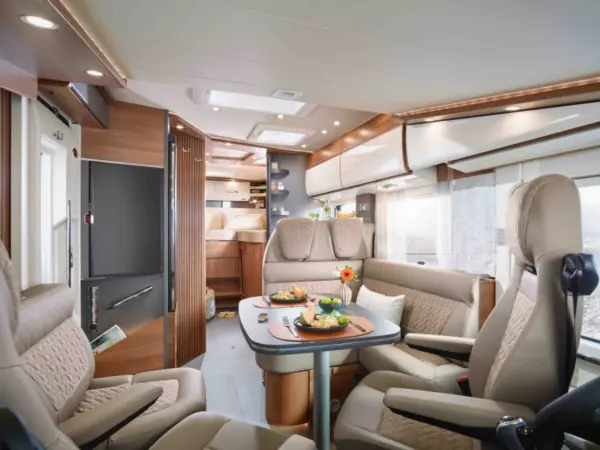Luxuriöses Carthago C2-Tourer Wohnmobil-Interieur mit beigen Ledersitzen, einem kleinen Esstisch mit Tellern mit Speisen und Getränken, Holzvertäfelung, Stauraum über Kopf und großen Fenstern, die natürliches Licht hereinlassen.