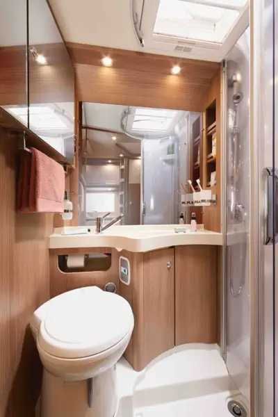 Ein modernes Carthago C2-Tourer Wohnmobil-Badezimmer mit Holzschränken, einer Toilette, einem Eckwaschbecken, einem Spiegel, Regalen für Toilettenartikel, einem Handtuchhalter und einer Glasduschkabine. Oberlichter und eine Dachluke erhellen den Raum.