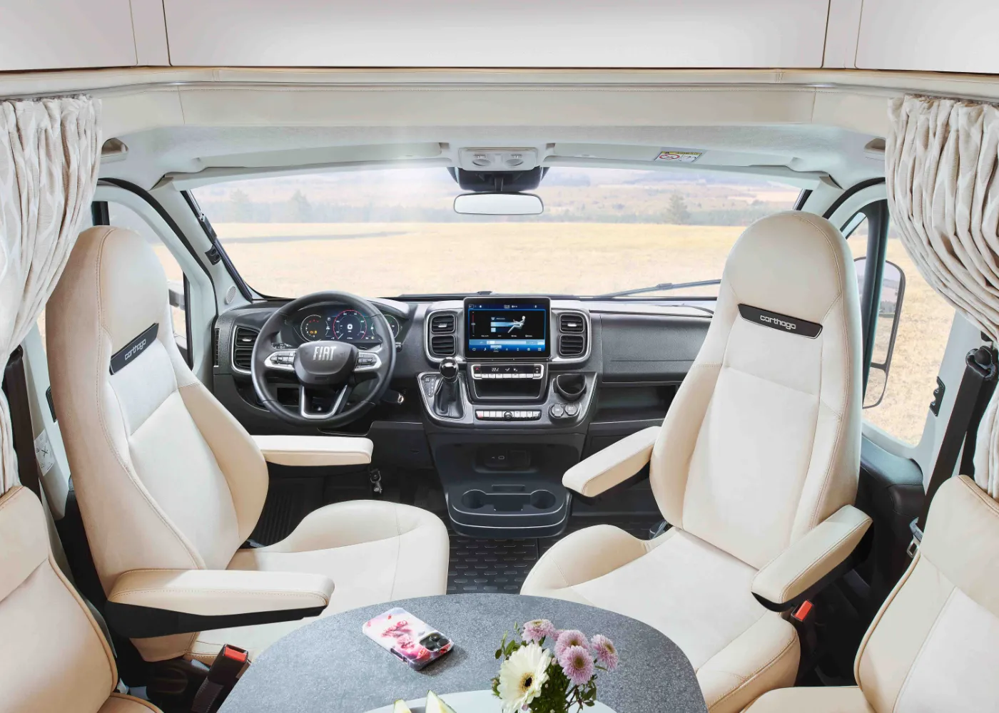 Innenansicht eines modernen integrierten Wohnmobil-Fahrerhauses mit zwei cremefarbenen Sitzen, Bildschirmen auf dem Armaturenbrett und einem kleinen Tisch mit einer Blumenvase und einem Smartphone. Durch die vordere Windschutzscheibe ist eine malerische Landschaft zu sehen.