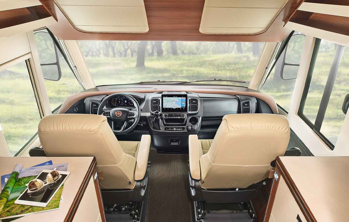 Innenansicht eines integrierten Wohnmobil-Cockpits mit zwei beigen Sitzen, einem Armaturenbrett mit eingebautem Navigationsbildschirm und großen Fenstern, die eine grasbewachsene Außenszene einrahmen. Im Vordergrund steht ein Tisch mit Prospekten und Snacks.