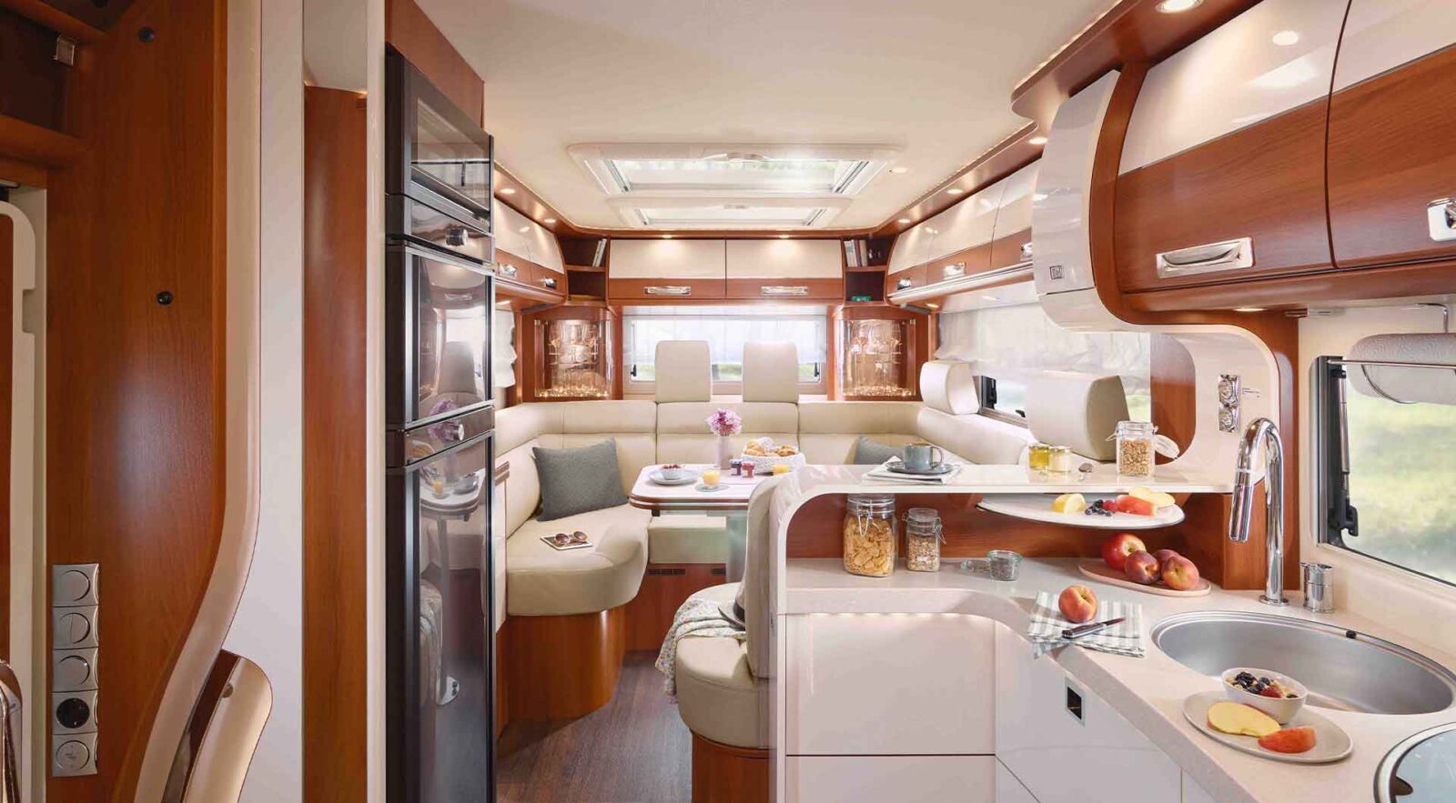 Modernes Wohnmobil-Interieur mit einer gemütlichen Essecke mit beigen Sitzgelegenheiten, einer kompakten Küche, Holzschränken und verschiedenen Lebensmitteln - dieses integrierte Wohnmobil kombiniert Funktionalität und Stil für komfortables Reisen.