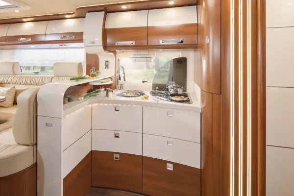 Die moderne Wohnmobilküche im Carthago Liner for Two verfügt über glänzend weiße und hölzerne Schränke, eine kleine Spüle, ein Kochfeld mit Pfannen und ein Fenster über der Theke. Der Raum ist durch eingebaute Lampen gut beleuchtet, und auf der linken Seite steht ein Sofa.