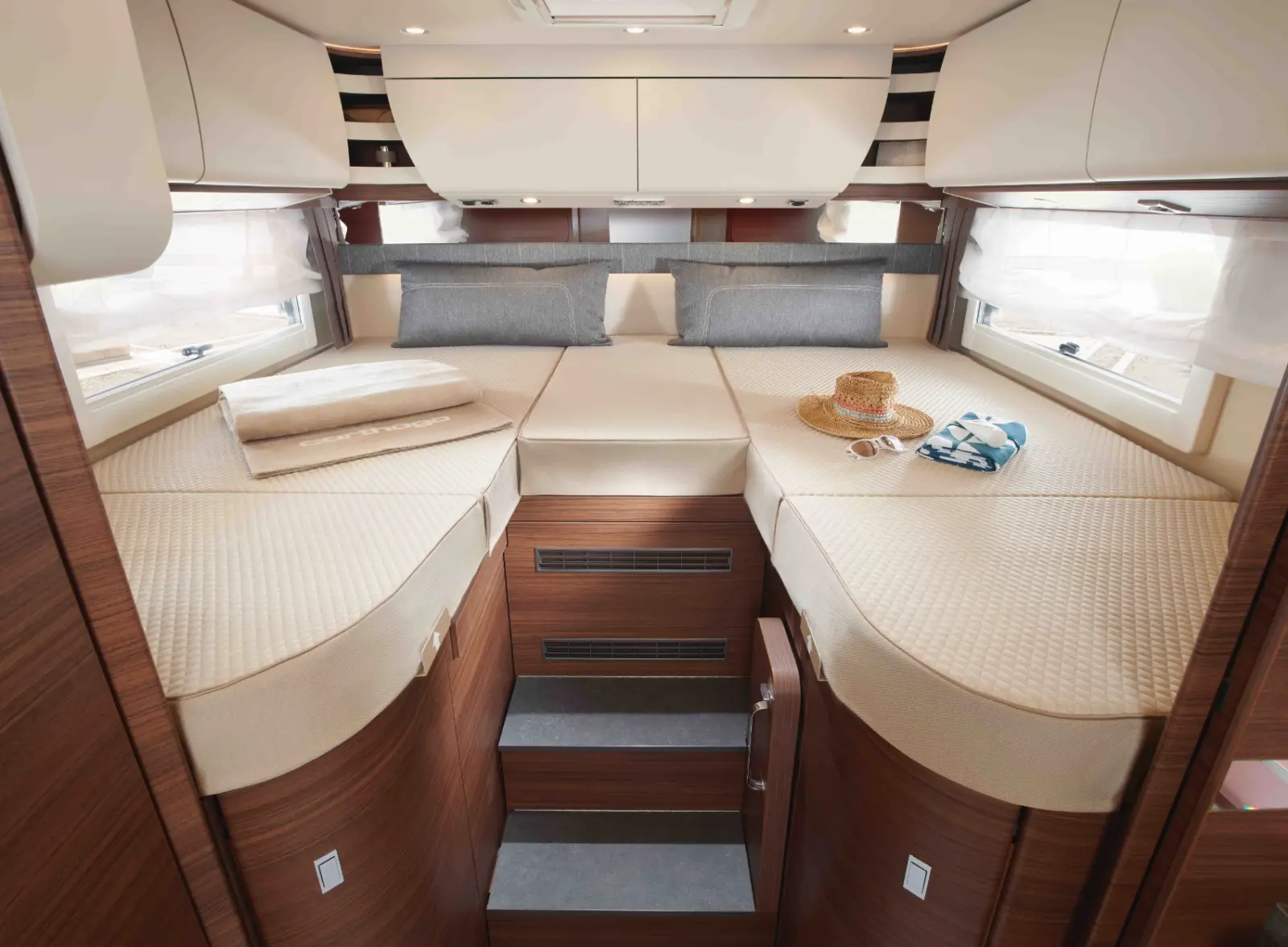 Interieur eines modernen teilintegrierten Wohnmobils mit zwei Einzelbetten und Bettzeug, durch Stufen getrennt; auf einem Bett liegen Strohhüte und ein gestreiftes Handtuch, darüber und darunter befinden sich Holzverkleidungen und Ablageschränke.