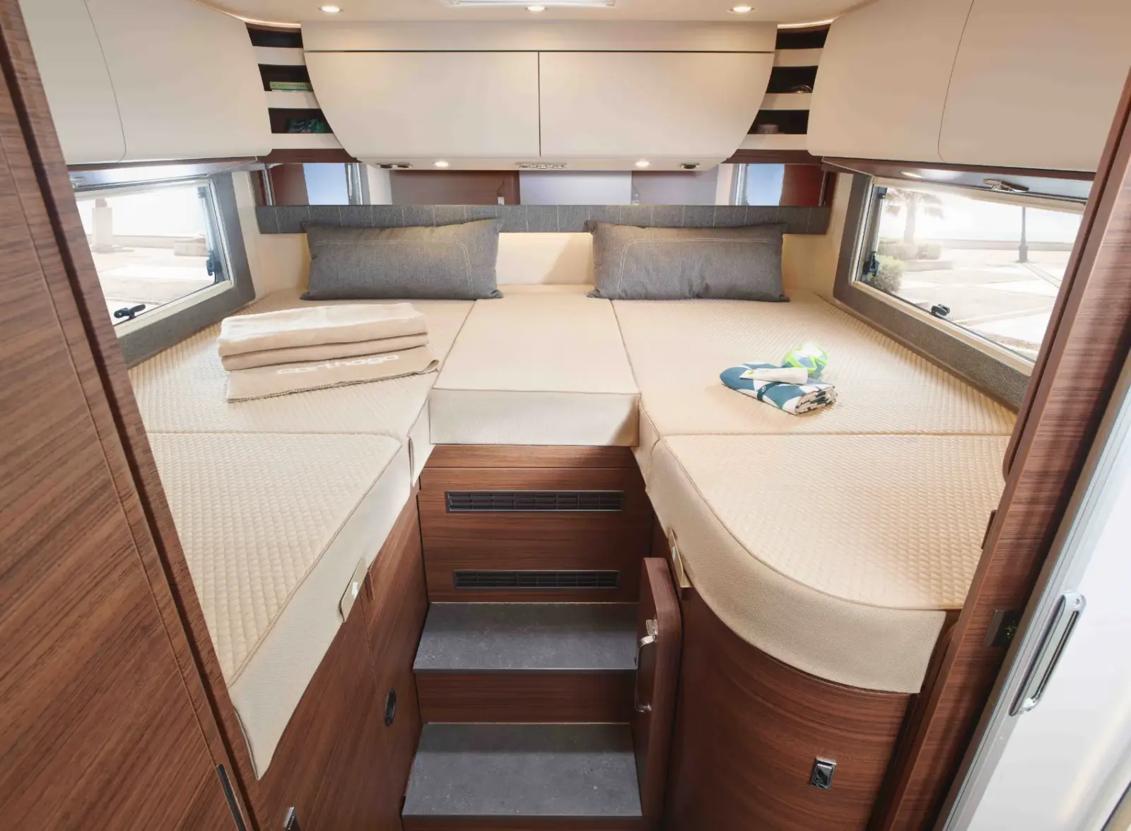 Interieur eines teilintegrierten Wohnmobil-Schlafzimmers mit zwei Einzelbetten, die zu einem großen Schlafbereich verbunden sind, brauner Holzvertäfelung, Stufen zwischen den Betten, gefalteten Handtüchern, grauen Kissen und Fenstern auf beiden Seiten.