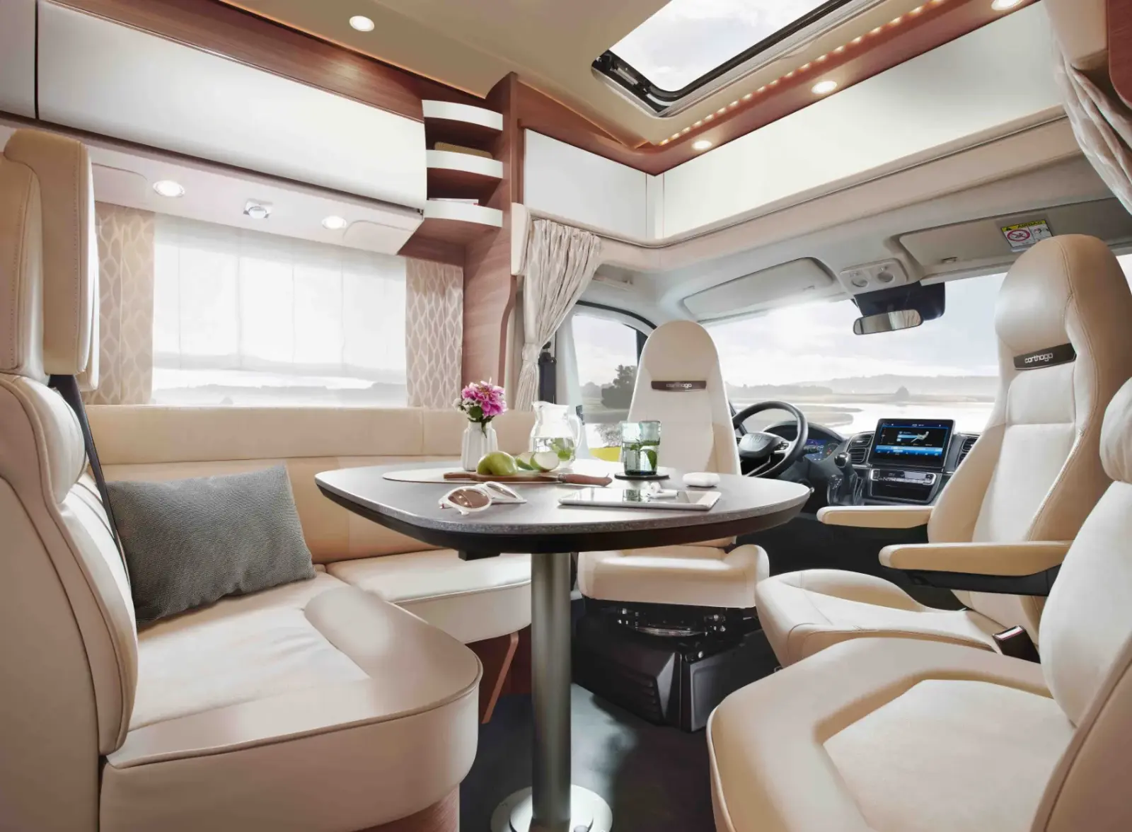 Modernes Wohnmobil-Interieur mit cremefarbenen Sitzen, einem runden Tisch mit Geschirr, Blumen und Getränken, großen Fenstern, die natürliches Licht hereinlassen, und einer Fahrerkabine im Hintergrund.