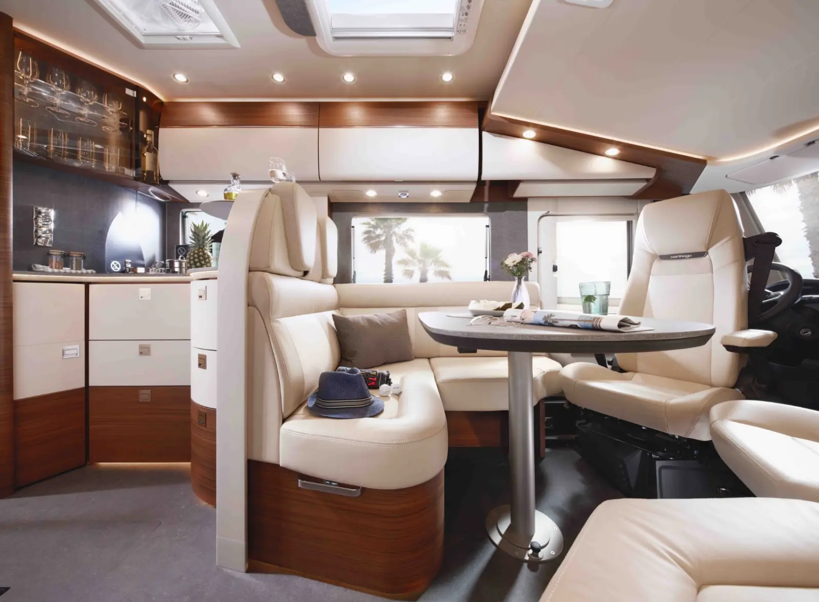 Modernes Wohnmobil-Interieur mit gepolsterten cremefarbenen Sitzen und hölzernen Akzenten, mit einem runden Tisch und einer kleinen Küche. Sonnenlicht strömt durch die Fenster und schafft eine gemütliche, einladende Atmosphäre.