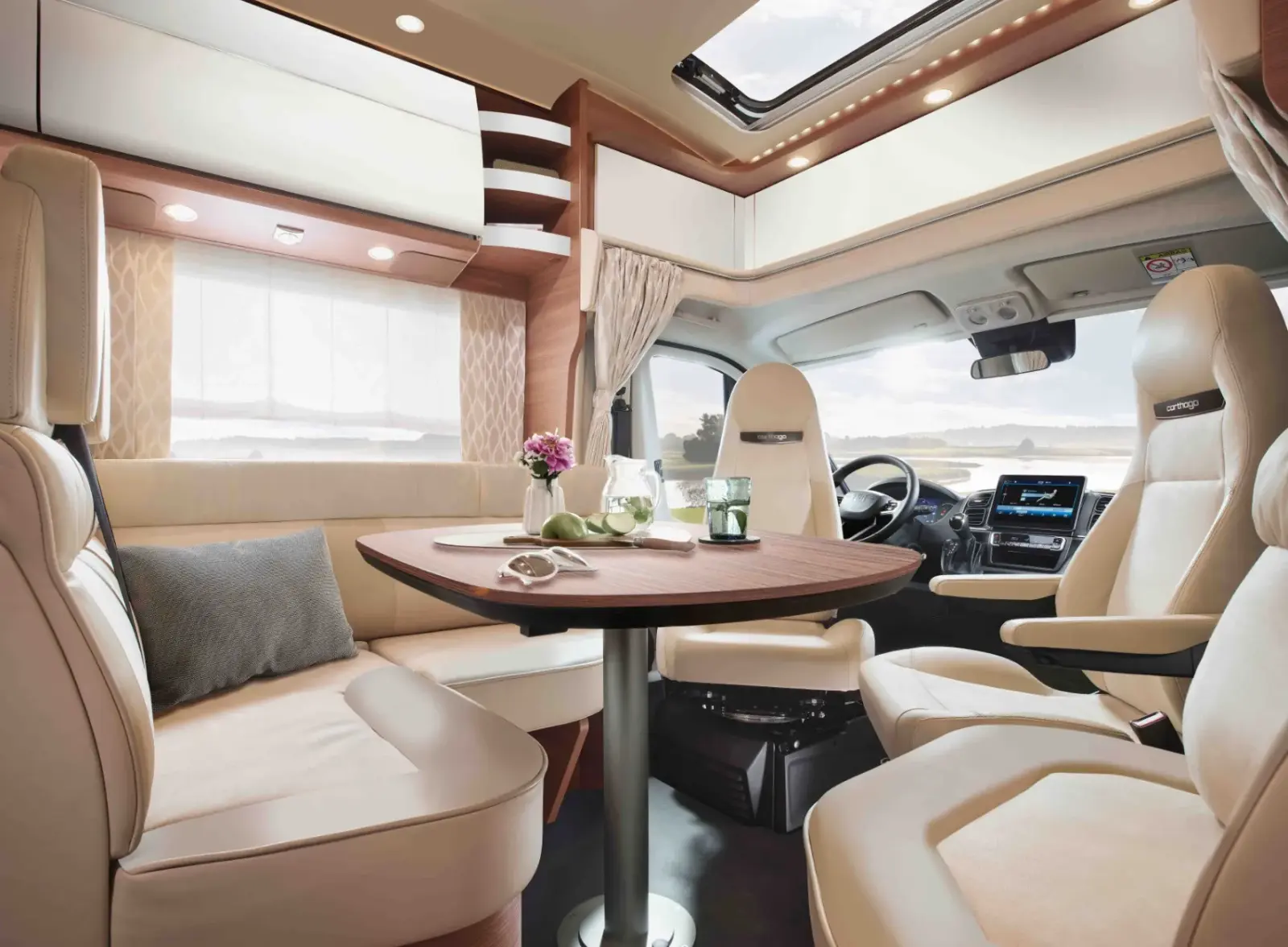 Modernes Wohnmobil-Interieur mit cremefarbenen Ledersitzen, einem runden Holztisch mit Getränken und Blumen, großen Fenstern mit Vorhängen und einem Schiebedach, das natürliches Licht hereinlässt. Im Hintergrund ist die Fahrerkabine zu sehen.