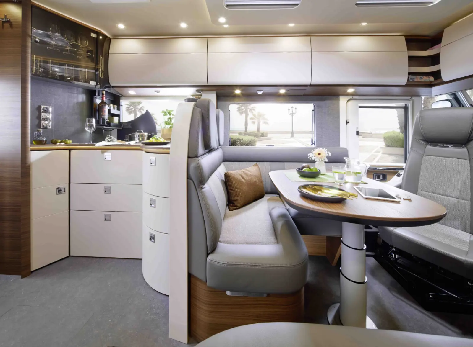 Modernes vollintegriertes Wohnmobil-Interieur mit geschwungenen Schränken, einem kleinen Küchenbereich und einer gemütlichen Essecke mit grauen und beigen Sitzgelegenheiten um einen Holztisch für mehrere Personen. Große Fenster sorgen für natürliches Licht und einen malerischen Blick nach draußen.