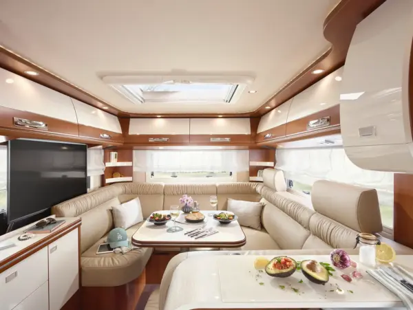 Das moderne Wohnmobil-Interieur des Carthago Liner for Two bietet beigefarbene Ledersitze, einen U-förmigen Essbereich, einen gedeckten Tisch mit Speisen und Getränken, Holzschränke, große Fenster mit Vorhängen und einen Fernseher für eine gemütliche, helle Atmosphäre.