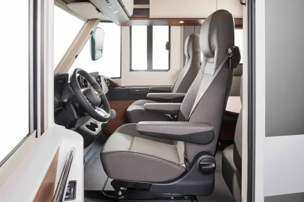 Innenansicht eines modernen Carthago Chic C Line Wohnmobils mit zwei Vordersitzen mit Armlehnen, einem Lenkrad, Getränkehaltern und großen Fenstern - alles in einem eleganten, farblosen Design.