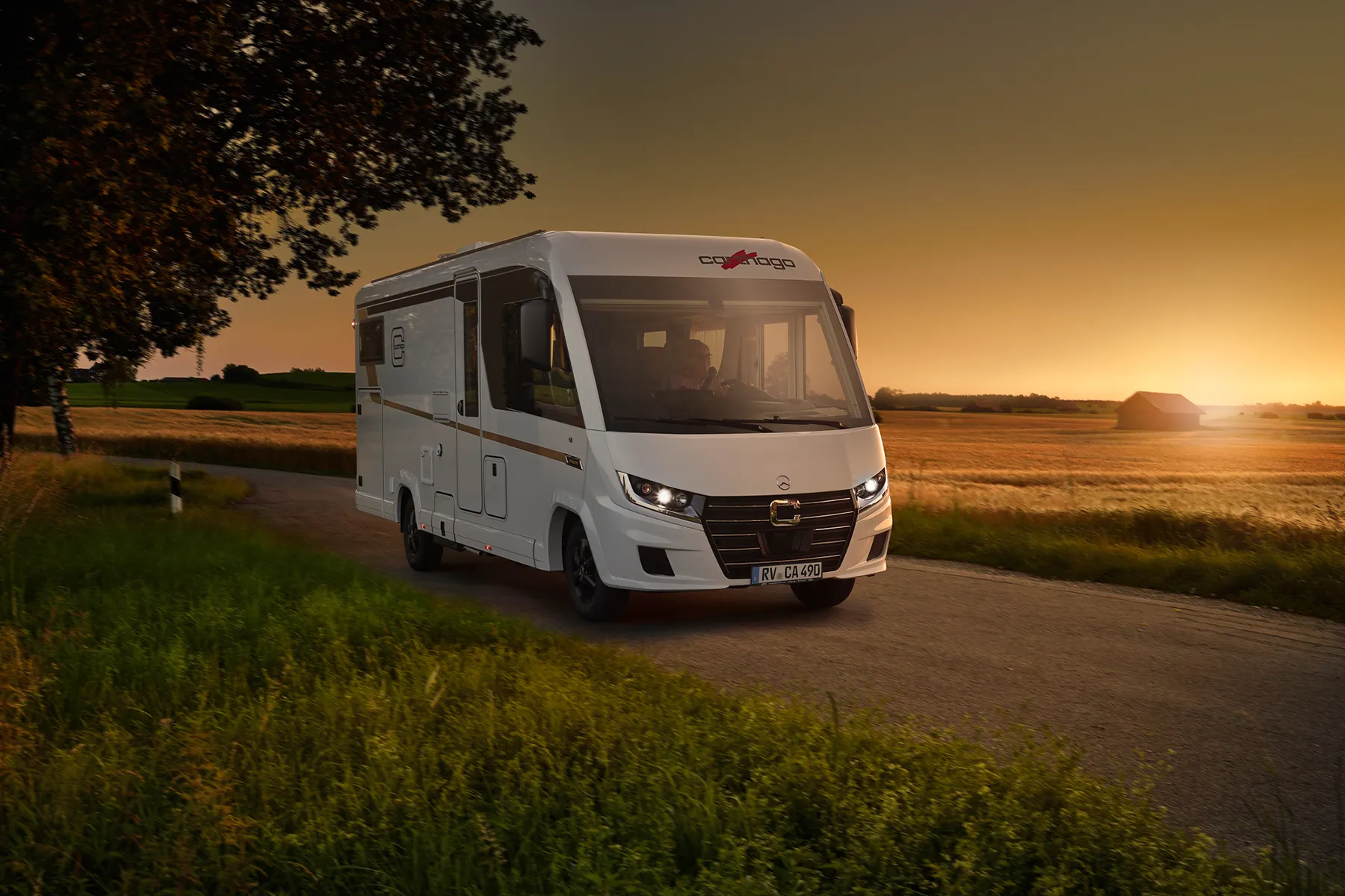 Ein Carthago Chic C-Line Wohnmobil fährt bei Sonnenuntergang eine Landstraße entlang, umgeben von grünem Gras, goldenen Feldern und weit entfernten landwirtschaftlichen Gebäuden unter einem orangefarbenen Himmel.