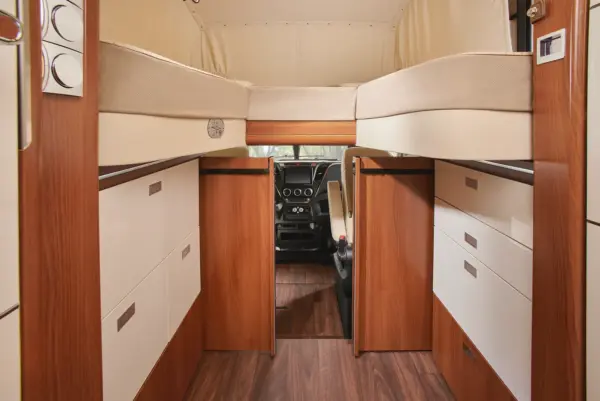 Innenraum eines Carthago Liner for Two Wohnmobils mit eleganten Holzschränken und Schubladen, einem erhöhten Bett darüber und dem Fahrer-Cockpit mit zwei schwarzen Sitzen am anderen Ende. Der moderne Raum bietet eine übersichtliche Aufteilung und einen stilvollen Fußboden in Holzoptik.
