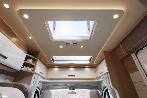 Die Innendecke eines modernen Carthago C Tourer Teilintegrierten Wohnmobils ist mit Holzschränken, eingebauten Leuchten und einem offenen Dachfenster ausgestattet, das Tageslicht hereinlässt. Die Oberseiten von zwei Sitzen und ein Teil der verglasten Tür sind sichtbar.