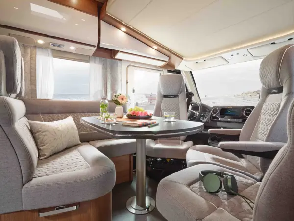 Das Innere eines modernen Carthago C2-Tourer Wohnmobils besteht aus grauen Plüschsitzen, die um einen mit Blumen, Obst, Getränken, einem Notebook und Kopfhörern gedeckten Tisch angeordnet sind, und großen Fenstern mit Blick auf den Strand.