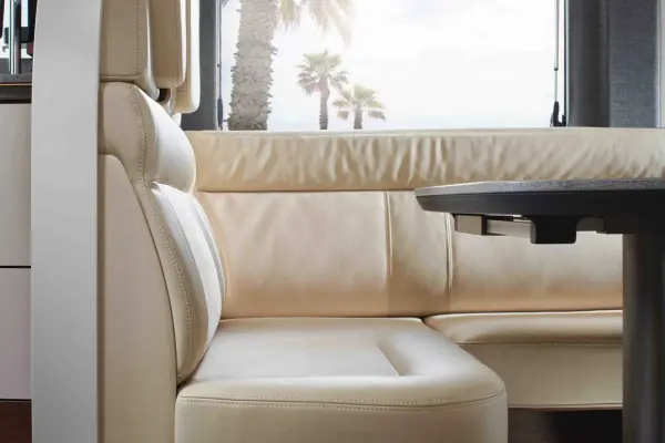 Eine Nahaufnahme eines beigen Ledersitzes und einer dunklen Tischplatte im Inneren eines Carthago Chic C-Line T Wohnmobils, mit Palmen und einer sonnigen Szene im Freien, die durch das Fenster im Hintergrund zu sehen ist.