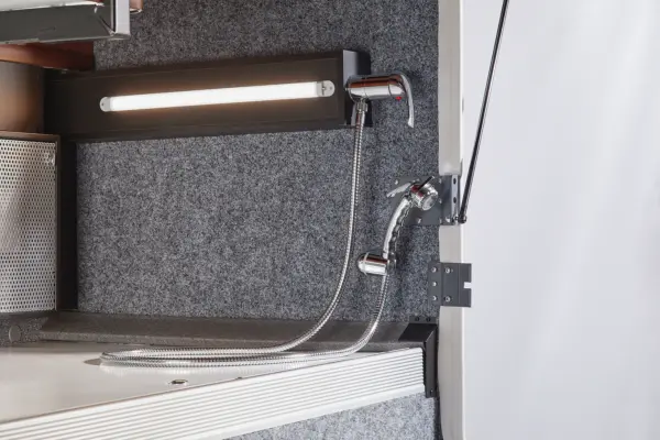 Eine Nahaufnahme eines kompakten, an der Wand montierten Wasserhahns und eines Duschkopfs mit flexiblem Metallschlauch in einem grauen, mit Teppichboden ausgelegten Carthago Liner for Two Transporter, neben einer horizontalen LED-Leuchte und einem Ablagekorb aus Metallgewebe.