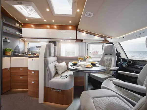 Modernes Wohnmobil-Interieur des Carthago C2-Tourer mit hellem Holz und cremefarbenen Möbeln, einem Esstisch für zwei Personen mit Speisen und Getränken, einer kompakten Küche und großen Fenstern, die den Blick auf das Meer freigeben. Der Raum wirkt hell und einladend.