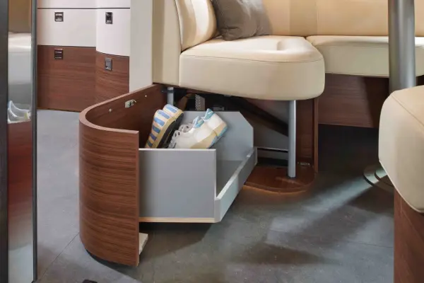 Eine geschwungene Holzschublade, die in ein beigefarbenes Carthago Chic C Line Sofa eingebaut ist, lässt sich öffnen und gibt den Blick auf einen Stauraum frei, in dem sich ein gestreifter Hut und weiße Schuhe befinden, die auf einem dunklen Boden stehen.