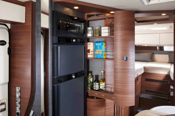 Die moderne Wohnmobilküche im Carthago Chic C-Line T verfügt über Holzschränke, eine offene Speisekammer mit Lebensmitteln und Gewürzen, eine eingebaute Mikrowelle und einen kleinen Kühlschrank neben einem ordentlich gemachten Bett im Schlafbereich.