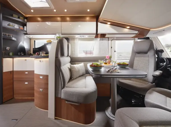 Modernes carthago c2-tourer Wohnmobil-Interieur mit geschwungenen Holzschränken, hellgrauen gepolsterten Sitzen um einen Esstisch mit Tellern mit Speisen und Getränken, großen Fenstern und hellem, natürlichem Licht.