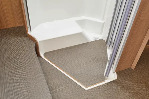 Eine Nahaufnahme eines modernen Duschbereichs im Carthago C2-Tourer mit einer weißen Duschwanne und einer braunen Bodenmatte, Wandpaneelen aus Holz und einer teilweise geöffneten Glastür.
