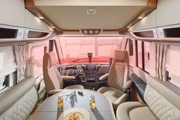 Blick ins Innere eines modernen Carthago Liner for Two mit Fahrer- und Beifahrersitz, einem runden Tisch mit zwei Getränken und Obst sowie großen Frontfenstern mit beigen Vorhängen und eingebauten Staufächern über dem Kopf.