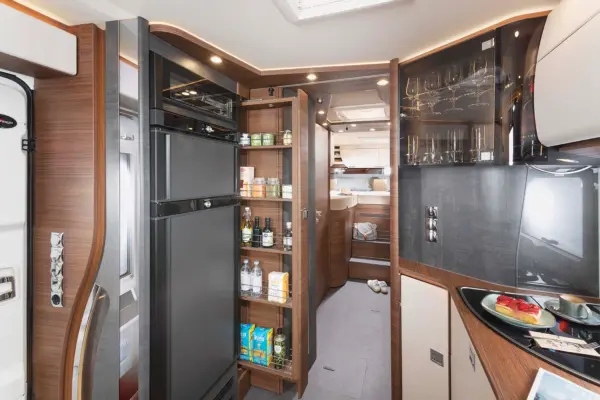 Moderne Carthago Chic C Line Wohnmobilküche mit einem hohen schwarzen Kühlschrank, einer offenen Speisekammer mit Regalen für Lebensmittel und Getränke, Gläsern in Schränken und einer Theke mit Snacks, die zu einem gemütlichen Schlafbereich im Hintergrund führt.