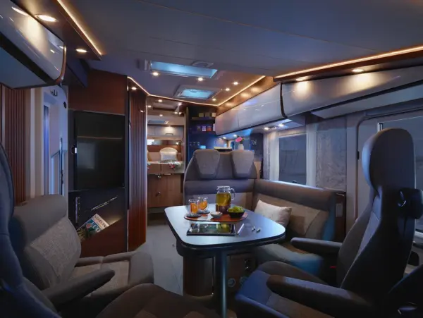 Modernes Wohnmobil-Interieur mit plüschigen grauen Sitzmöbeln um einen Tisch für Tee, Gläser und Snacks. Warme Ambientebeleuchtung, Holzakzente und Regale schaffen eine gemütliche Atmosphäre in dieser Küche und dem Wohnbereich des Carthago C2-Tourer.