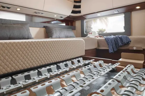 Ein modernes Wohnmobil-Schlafzimmer in einem Carthago Chic C Line mit einem erhöhten Bett, das Stauraum unter dem Bett bietet, mit einem freiliegenden Betthebe-Mechanismus, neutraler Bettwäsche, zwei grauen Kissen und einem Fenster, das die Palmen draußen einrahmt.
