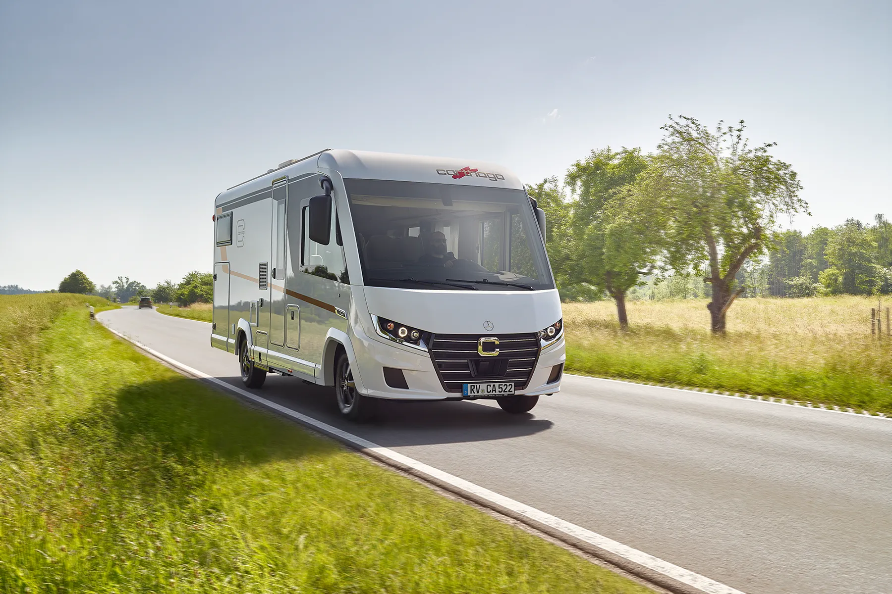 Ein Carthago Chic C Line Reisemobil fährt auf einer Landstraße, umgeben von grünem Gras und Bäumen, unter einem klaren blauen Himmel.